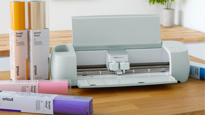 Light blue Cricut Explore machine on a table with multiple tubes of colored vinyl on the table next to it